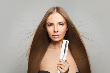 Studio portrait of fashion model woman using hair straighteners iron on white background. Harstyle, hair care, beauty salon and treatment conceptの写真素材