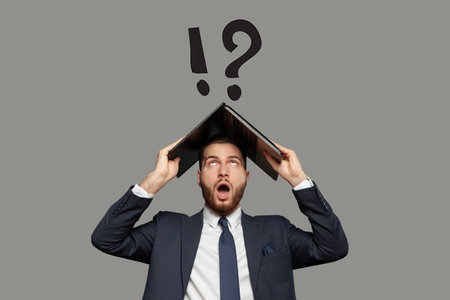 Emotional shocked businessman in suit with laptop on his head on gray background with question and exclamation marks. Disapointment conceptの写真素材