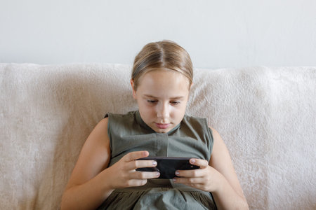 Young girl using the phone, playing the game, sitting on sofa at cozy home. Child and smartphone. Technology, communication and leisure activity conceptの写真素材