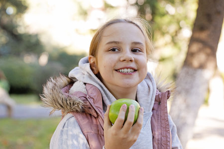 Smiling kid holding green apple. Young girl in park, outdoors portraitの写真素材