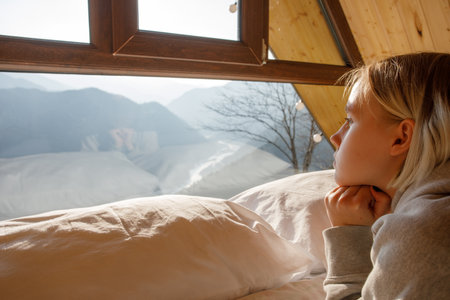 Pensive blonde woman laying on the bed and looking at the mountains in the windowの写真素材