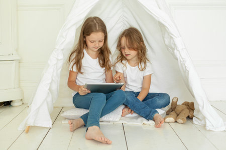 Playful little girl sisters with tablet pc computer in kids tent at home. Childhood, technology and hygge conceptの写真素材