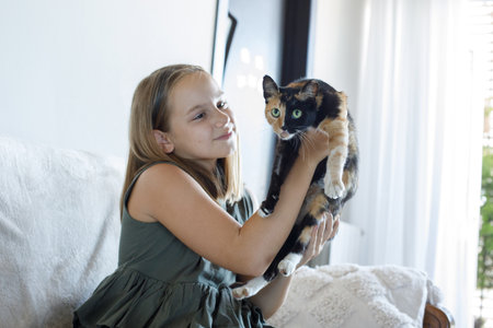 Adorable young girl hugging her cat pet sitting on cozy sofa, relaxing at home. Child and domestic kitten indoor. Love, bonding and domestic life conceptの写真素材