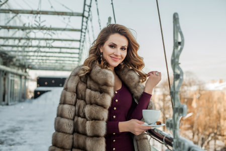 Smiling happy brunette woman wearing fur coat holding a cup of coffeeの写真素材