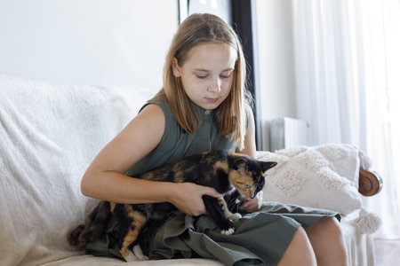 Cheerful young girl hugging her cat pet sitting on cozy sofa, relaxing at home. Child and domestic kitten indoor. Love, bonding and life conceptの写真素材