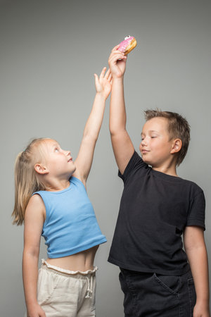 Playful sister and brother having fun holding donuts standing against white background wall. Child and chocolate glazed donut indoorの写真素材