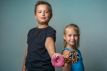 Beautiful sister and brother having fun holding donuts standing against blue background wall. Child and chocolate glazed donut indoor.の写真素材