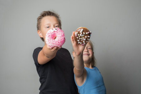 Attractive sister and brother having fun holding donuts standing against white background wall. Child and chocolate glazed donut indoorの写真素材
