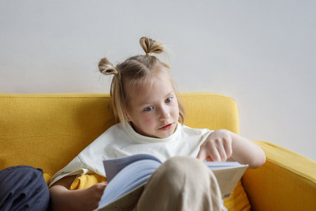 Portrait of little smart child girl looking at book while sitting on sofa indoorの写真素材