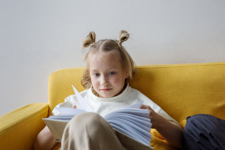 Cheerful little girl sitting on sofa reading book at home. Childhood and people concept