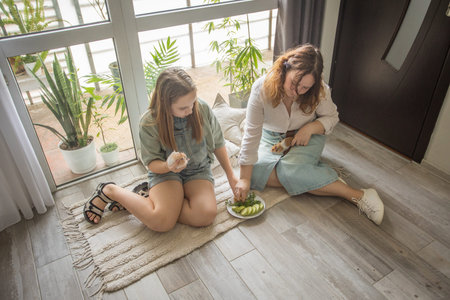 girl and women relax on a rug in a modern indoor space. They share snacks while sitting on a wooden...
