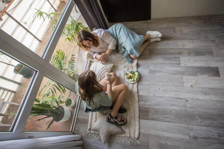 girl and women relax on a rug in a modern indoor space. They share snacks while sitting on a wooden...