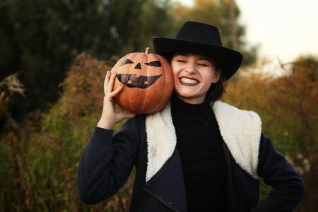 Girl in a black hat holds a pumpkin on his shoulder and laughs closing her eyesの写真素材