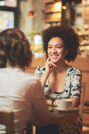 Mixed race woman in coffee shop having coffee with friend and smilingの写真素材