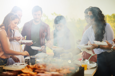 Group of people standing around grill, chatting, drinking and eating.の写真素材