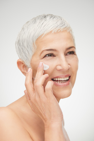 Head shot of beautiful senior woman putting on the cream.の写真素材