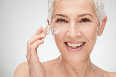 Head shot of beautiful senior woman putting on the cream.の写真素材