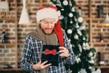 Surprised Caucasian man standing in front of Christmas tree and opening gift. Santa's hat on the head.の写真素材