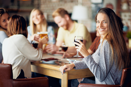 Woman looking at camera and holding glass of wine while sitting in restaurant. In background friends drinking and chatting.の写真素材