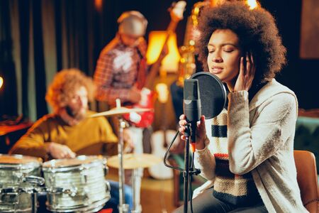 Mixed race woman singing. In background band playing instruments. Home studio interior.の写真素材