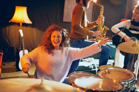 Talented female drummer playing drums. In background the rest on the band playing instruments. Home...