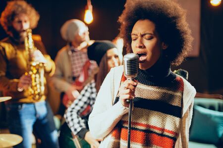 Close up of mixed race woman singing. In background band playing instruments. Home studio interior.の写真素材