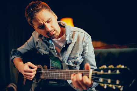 Caucasian talented bearded musician playing his acoustic guitar and sitting on sofa. Living room...