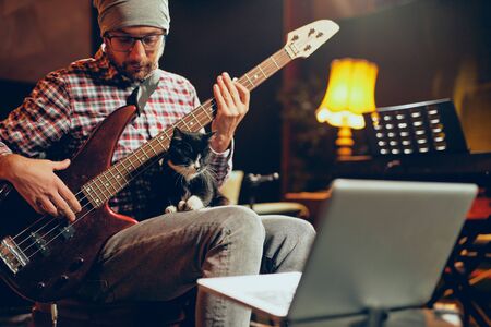 Young Caucasian man with hat on head playing bass guitar while sitting in home studio.