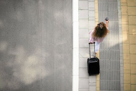 Top view of woman carrying the luggage and holding coffee to go while walking to the station.