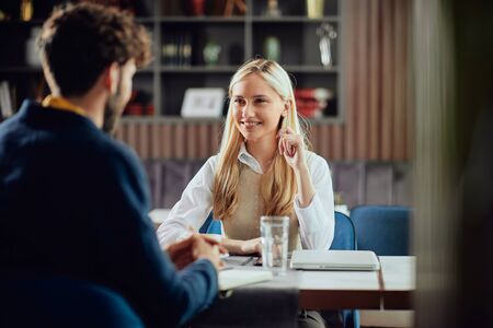 Smiling Caucasian blonde businesswoman dressed smart casual discussing with her male colleague about project while sitting in coffe shop.の写真素材