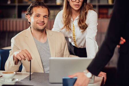 Arrtactive businessman dressed smart casual sitting in cafe and looking at camera. Around him his...