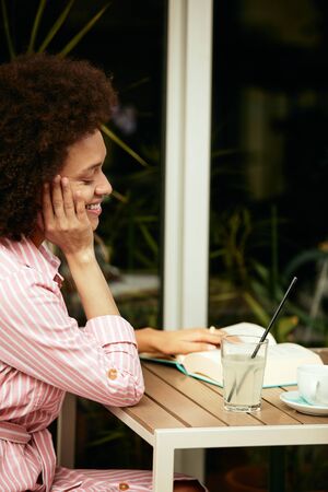 Attractive mixed race woman in pink striped dress reading book in cafe. On table are coffee and...