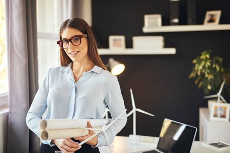 Attractive smiling businesswoman in formal wear and with eyeglasses standing in modern office and holding blueprints and windmill model. Sustainable development concept.の写真素材