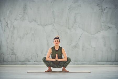 Front view of beautiful Caucasian brunette doing Squatting Balance yoga pose barefoot on mat.