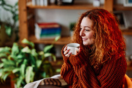 A cute ginger girl with curly hair is sitting in the chair at home in the morning and drinking her coffee.の写真素材