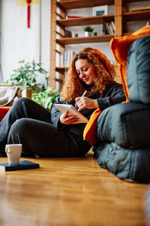 A cute ginger girl with curly hair is sitting in the chair at home in the morning and drinking her...