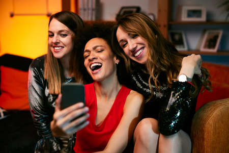 Three multicultural female friends sit in the living room in the evening and take selfies at home party.の写真素材