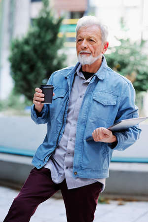 A happy senior student is standing on the street and holding his laptop and cup of coffee. Senior...