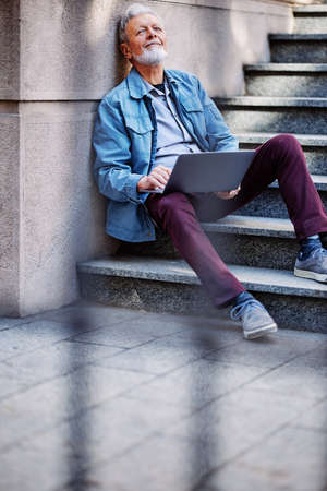 A senior student sits on the stairs of the university building and uses his laptop to look at notes...