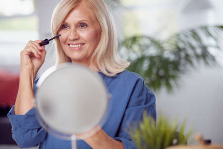 A happy senior woman sitting at home and putting mascara on eyelashes. Senior woman using make-upの写真素材