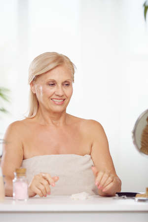 A senior blond woman is sitting at home surrounded by beauty products. Beauty and seniors, makeup for senior womanの写真素材
