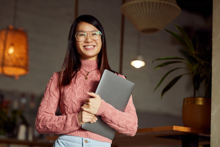 A happy Asian female student sitting in a cafe and posing with a laptop in her hands.の写真素材
