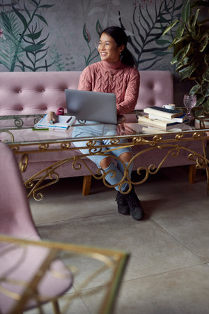 A happy Asian female student is sitting in the cafeteria and typing a report on her laptop. There are books on a table. Student exchange.の写真素材