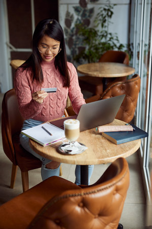 An Asian girl sitting in the cafeteria and using her credit card for online shopping on the laptop.の写真素材