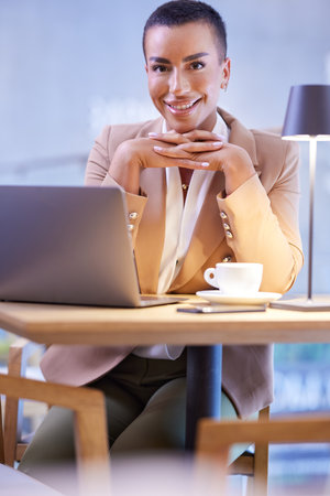 A friendly casually dressed businesswoman with short hair sits in a cafeteria and smiles at the camera. There is a laptop on a table.の写真素材