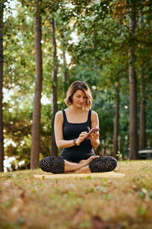 A slim yogi woman sits in a forest in lotus pose and uses phone.の写真素材