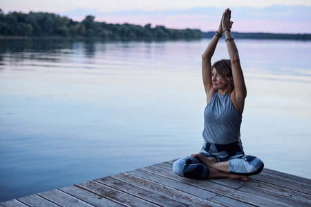 A calm woman in the lotus pose sits on a dock and practices yoga breathing near the river.の写真素材