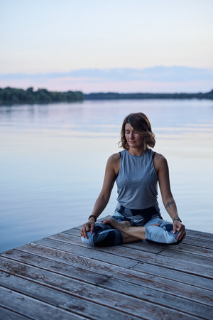 A calm woman in the lotus pose sits on a dock and practices yoga breathing near the river.の写真素材