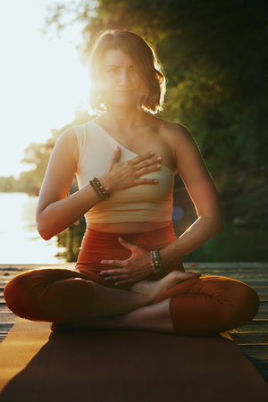 A calm woman in the lotus pose sits on a dock and practices yoga breathing near the river.の写真素材