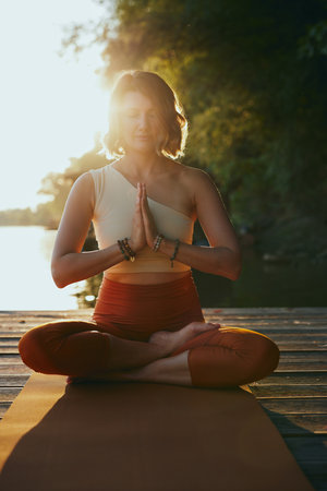 A calm woman in the lotus pose sits on a dock and practices yoga breathing near the river.の写真素材